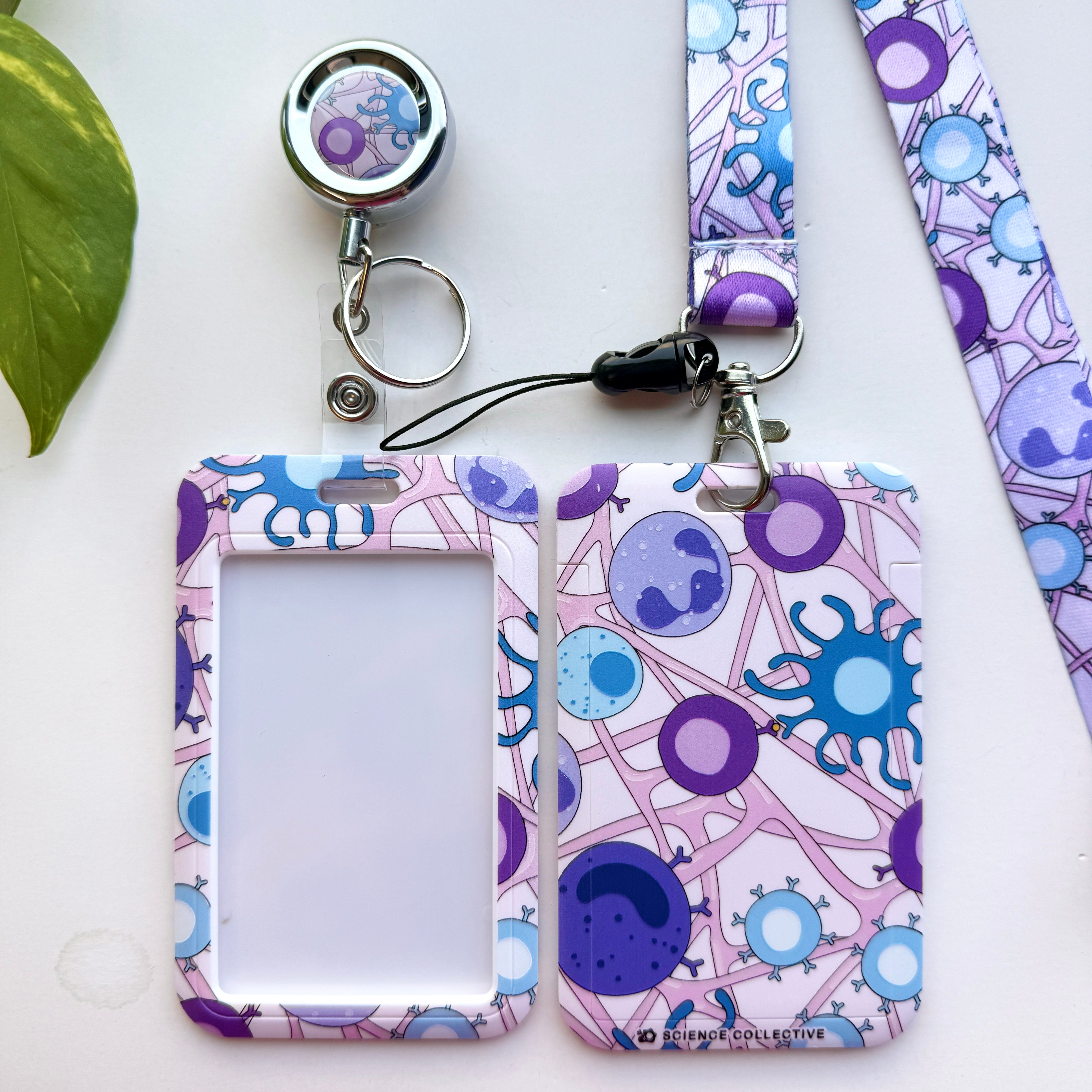 A flat-lay product shot of a matching ID immunology badge holder set with a retractable badge reel, a lanyard, and two card sleeves featuring a purple, blue, and white immune cells pattern.